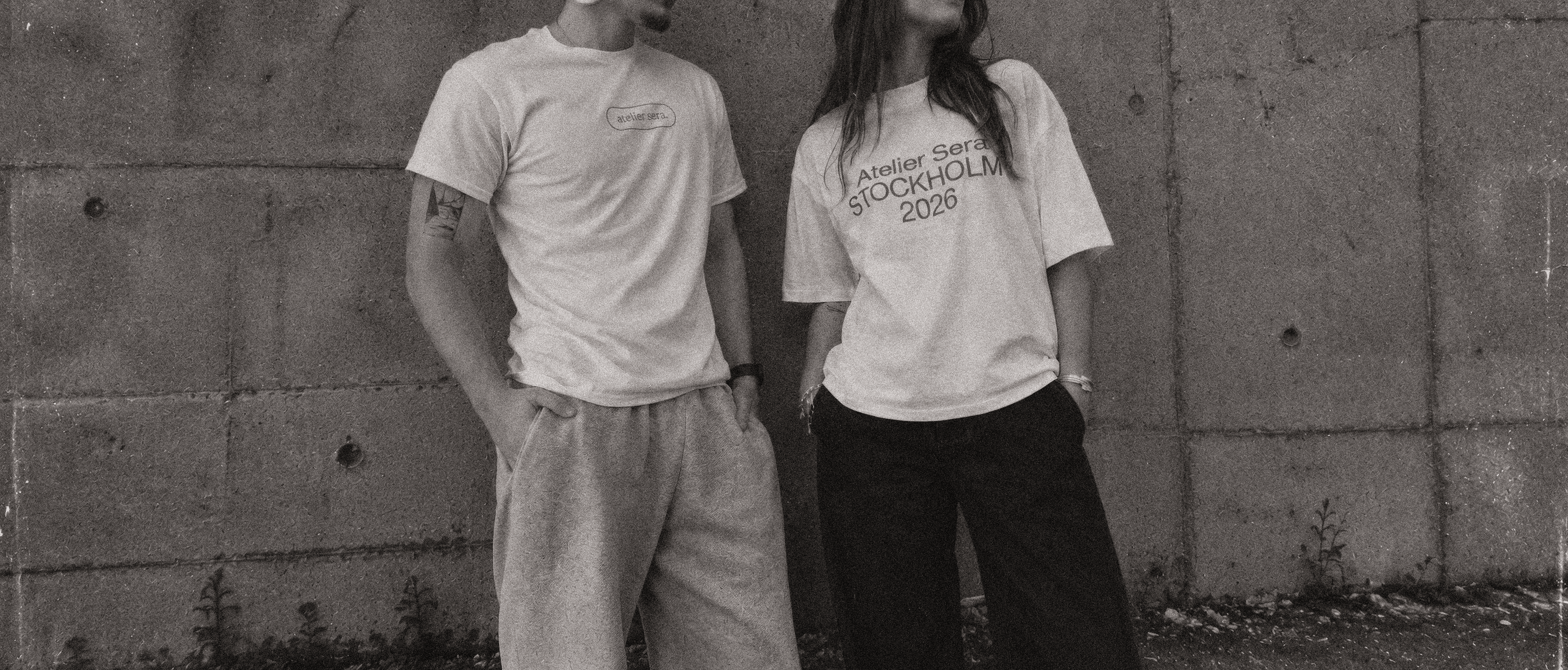 Two people wearing Atelier Sera tees against a concrete wall.